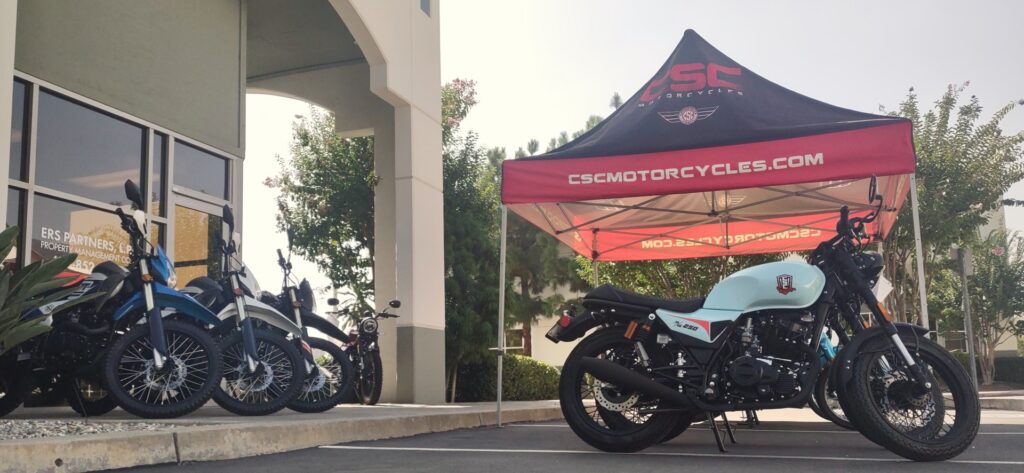 SG250 and TT250 Motorcycles parked in front of CSC Motorcycles under a pop-up tent.
